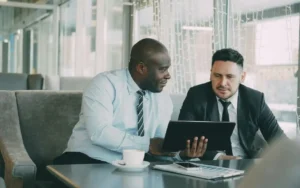 Two colleagues in business attire sit on a sofa in a bright lounge, examining a tablet together over a coffee break.