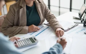 Two colleagues review financial documents at a bright desk, one pointing to a chart while the other uses a calculator,
