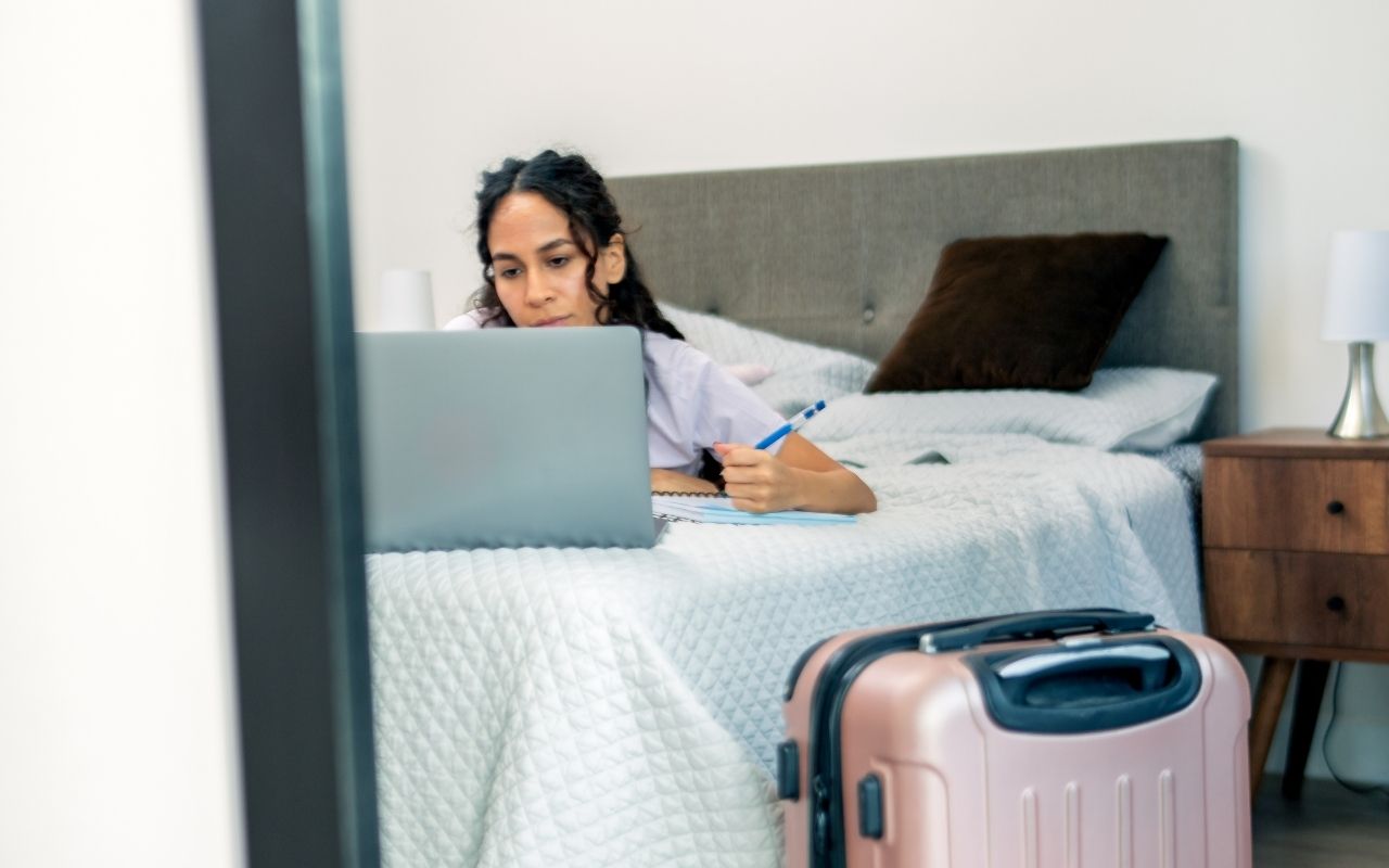 A woman lying on a bed in a rental room working on a laptop with a pink suitcase in the foreground, suggesting a long-term travel stay.