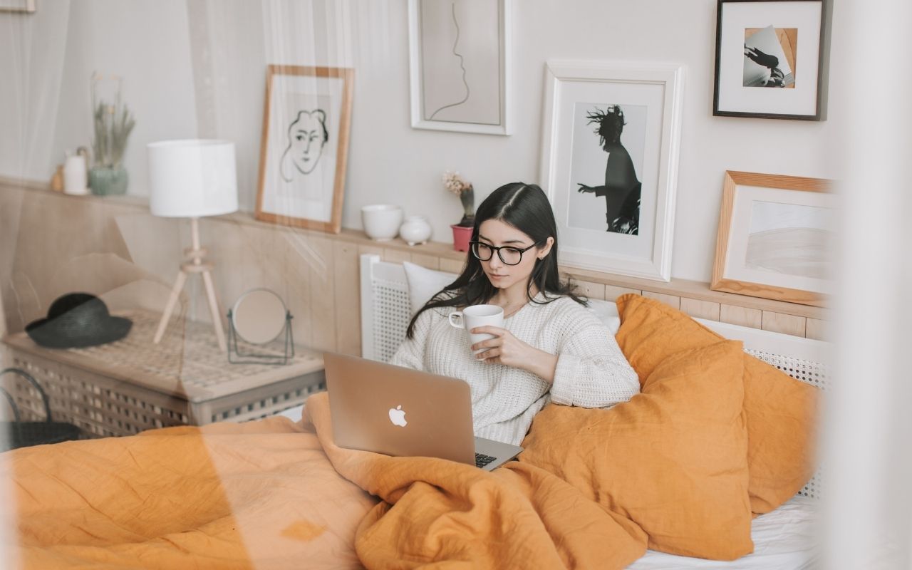 Woman with glasses sits in bed under an orange blanket, using a laptop and holding a mug.
