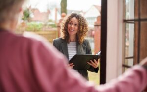Professional woman with curly hair speaks to someone at a doorway, holding a black binder/folder outside a house.