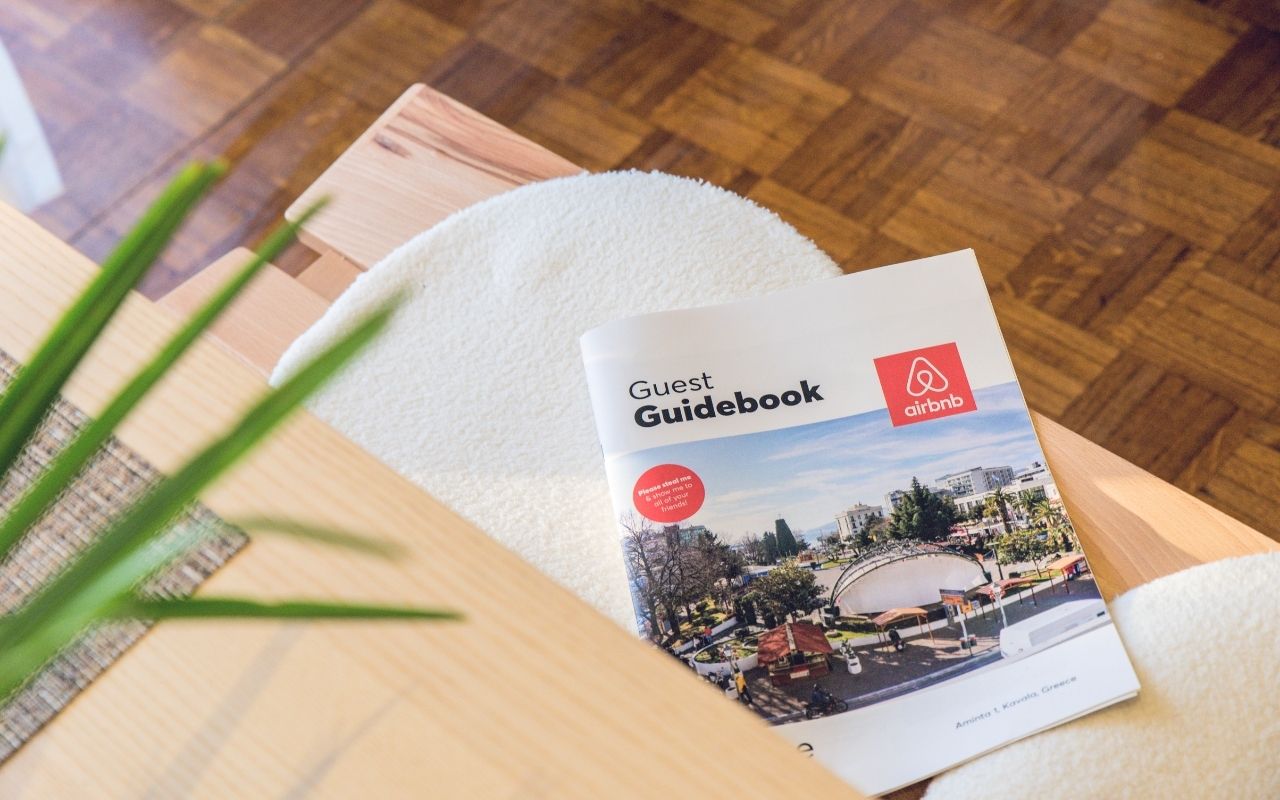 A close-up, high-angle shot of a printed Airbnb Guest Guidebook resting on a light wood coffee table with a white textured cushion. The guidebook features a scenic city photo on the cover and the red Airbnb logo in the top right corner.